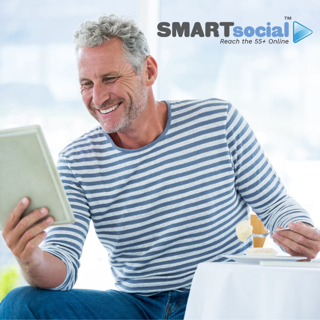55 YEAR OLD MAN EATING WHILE READING TABLET AND SMILING